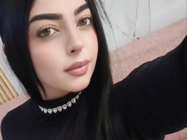 LilithAmoth, met zwart haar, bruine ogen en een neusring, draagt een zwarte top met een diamanten ketting. Ze heeft roze lippen en een subtiele glimlach en maakt een selfie..