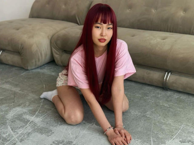 Yasmina, cheveux longs et roux, t-shirt rose, chaussettes blanches et short beige, agenouillée sur un tapis gris, regardant la caméra, avec un canapé vert derrière elle..