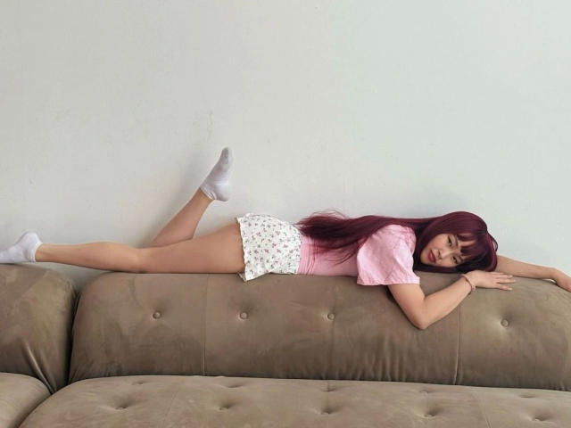 Yasmina: capelli rossi lunghi, top rosa, pantaloncini bianchi con fiori, calze bianche, sdraiata a faccia in giù su un divano beige, gambe sollevate, braccia tese in avanti..