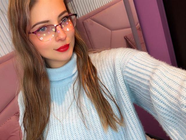 AmbeerWood, avec ses longs cheveux bruns, son rouge à lèvres rouge, ses lunettes et son pull en tricot bleu clair, se montre dans un autoportrait où elle met en valeur sa poitrine..
