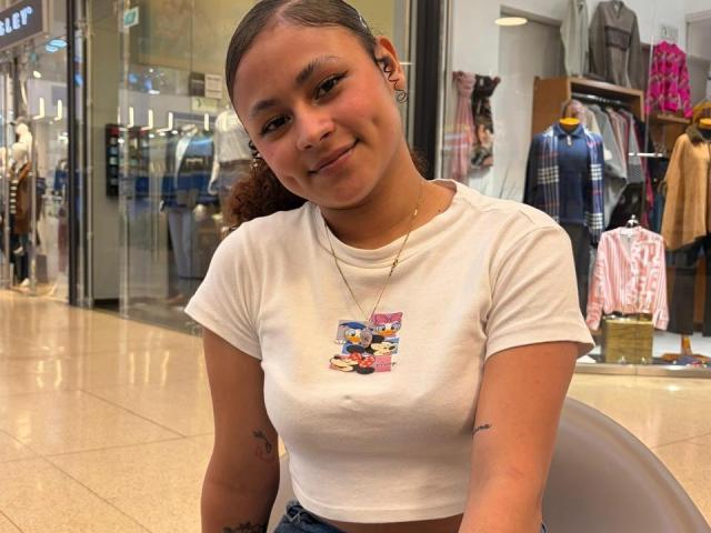 SofiaPassionne, una ragazza carina con la pelle marrone e piccoli seni, indossa una maglietta bianca con la stampa di Elmo, sorride, ha le cuffie nelle orecchie, piccoli tatuaggi sulle braccia, è in piedi in un centro commerciale e guarda la fotocamera..