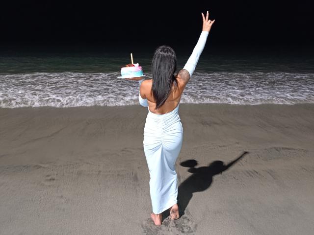NenaBlack, con lunghi capelli neri, indossa un abito bianco senza spalline e si trova su una spiaggia, tenendo una torta, con un braccio alzato, la sua ombra sulla sabbia, l'oceano sullo sfondo..