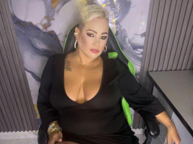 GianellaCox with big tits, black dress, blonde updo, green chair, gold earrings, bracelets, tattoo on right shoulder, pink lipstick, sitting, looking at camera, indoors.