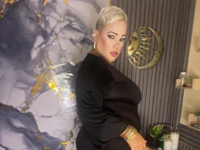 GianellaCox with short platinum blonde hair, wearing a black top, looking back over her shoulder, showing her large tits, standing in a modern room with a gold sun wall.