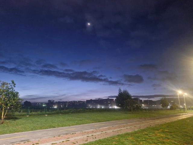 Céu noturno com uma lua, nuvens azul-escuras, campo gramado, rua com luzes e uma fileira de árvores ao fundo. As nádegas de BrendaCantorel não estão visíveis..