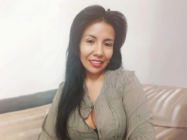 HeisseGinny, attraente con lunghi capelli neri, sorridente, indossa una camicia verde con bottoni, mostrando un po' di scollatura nera, seduta su un divano in pelle beige..