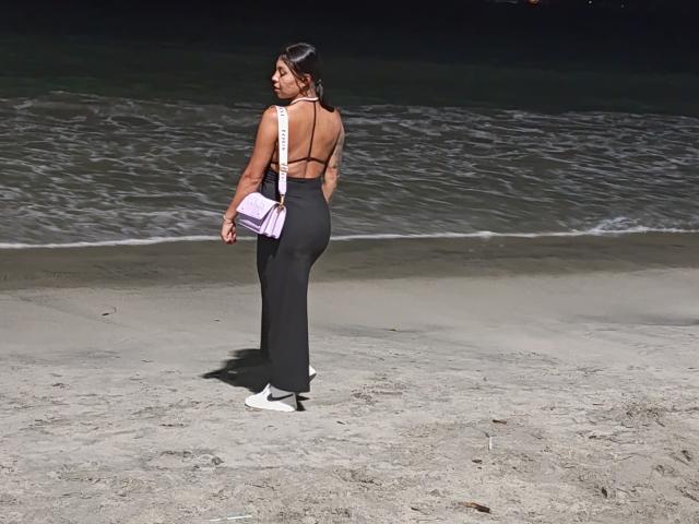 NenaBlack è in piedi su una spiaggia sabbiosa, con la schiena rivolta, indossa un abito nero senza schienale, una borsa rosa, scarpe da ginnastica bianche e mostra una linea di abbronzatura sui glutei..