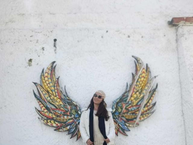 TheresaLarson with colorful, bird-like wings on a white wall, wearing a beige headscarf, white jacket, and black vest, standing with a calm, slightly serious expression.