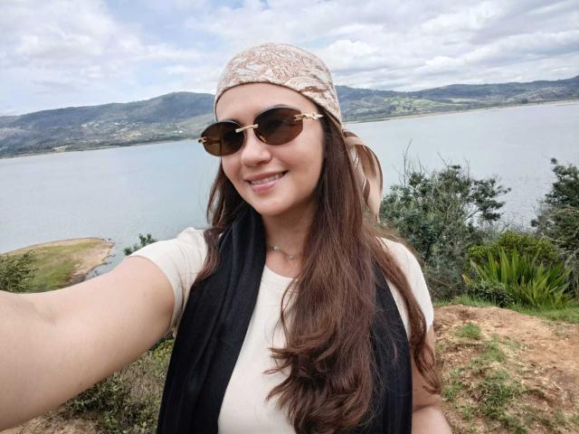TheresaLarson with long brown hair, wearing a beige patterned headscarf, black scarf, white shirt, and brown aviator sunglasses, smiling at the camera, taking a selfie by a lake.