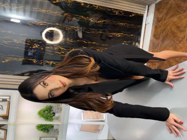 MilyEvans with long brown hair, black blazer, and black top, leaning forward, looking at camera, showing her tits, on a white surface, with a ring light in background.