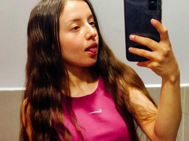 VioletaNova met lang bruin haar, een roze Nike sporttop, die haar tong uitsteekt, een telefoon vasthoudt en een selfie maakt, met een gemiddelde borstmaat, lichte huid, reflectie in een spiegel..