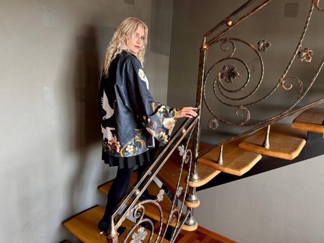 RubyDavis with curly blonde hair, wearing a black floral dress, standing on a wooden staircase with ornate metal railing, looking back over her shoulder.