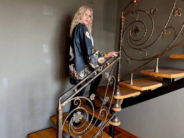RubyDavis with blonde curly hair, wearing a dark blue floral shirt, standing on a wooden staircase with ornate metal railings, looking back over her shoulder, hands on the railing.