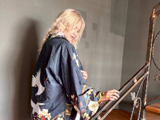 RubyDavis with blonde wavy hair, wearing a black floral kimono, stands on a metal staircase, looking over her shoulder, holding the railing, in a softly lit room.