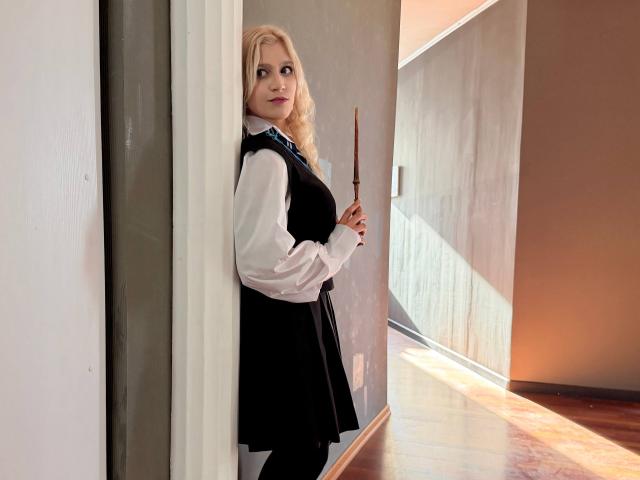 RubyDavis with blonde hair, wearing a black dress over a white shirt, holding a wooden stick, standing in a sunlit hallway, looking over her shoulder with a slight smile.