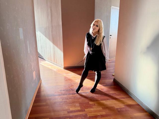 RubyDavis with blonde hair, black dress, white blouse, black tights, and black boots, standing in a sunlit hallway, leaning forward, showing her ass.