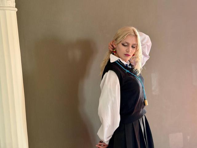RubyDavis with blonde hair, white shirt, black vest, and skirt, hand behind head, looking down, shadow on wall, blue necklace, standing, soft light, slightly smoky background.