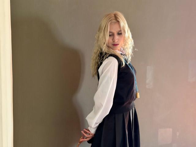 RubyDavis with blonde wavy hair, wearing a black sleeveless top over a white shirt, black skirt, and blue tie, standing against a wall, looking down with a slight smile.
