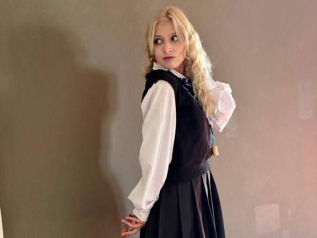 RubyDavis with blonde hair, wearing a white shirt, black vest, and black skirt, standing with a hand on her hip, looking back with a slight smile, tits visible.