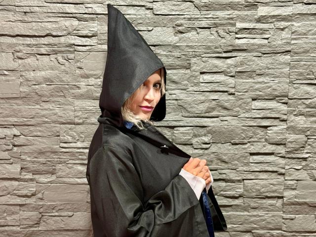 RubyDavis wearing a black hooded raincoat with a pointed hat, holding the coat open to show a white shirt. She has blonde hair, pink lipstick, and a cute smile.