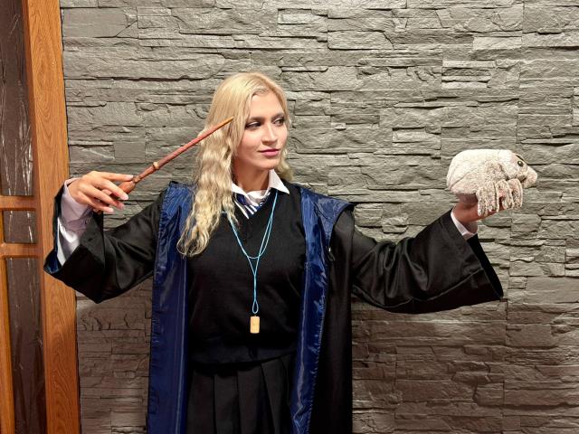 RubyDavis with blonde hair, wearing a black robe and white shirt, holding a wand in one hand and a rock-like object in the other. Stone wall in background.