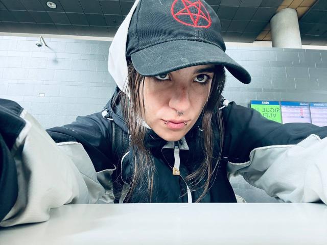 AmySpider with a black and white baseball cap, pentagram logo, nose ring, lip ring, and long brown hair, wearing a black and gray jacket, leaning forward.
