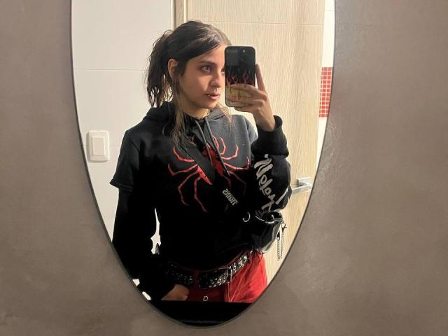 AmySpider with wet dark hair, black spider-print hoodie, red pants, holding phone in oval mirror selfie, showing slight pout, phone case has anime-style design, standing with hands in pockets.