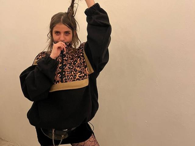 AmySpider with long brown hair, wearing a black oversized sweater with leopard print and beige stripe, holding her hair up, covering her mouth, in fishnet pants.