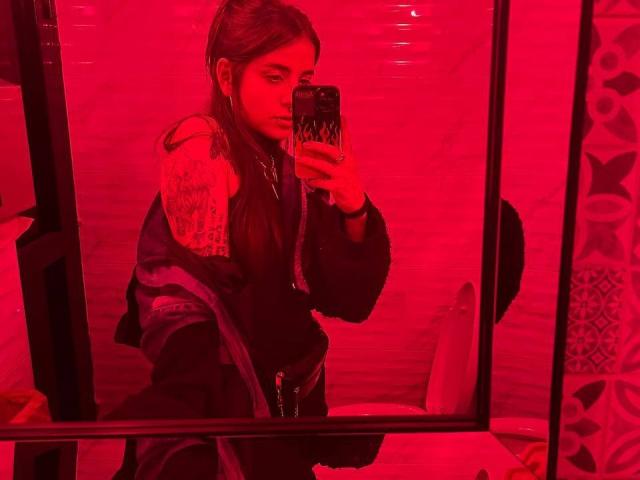 AmySpider with long brown hair, black top, and black jacket, holding phone in red-lit mirror, showing a serious face, her ass slightly visible under the jacket.