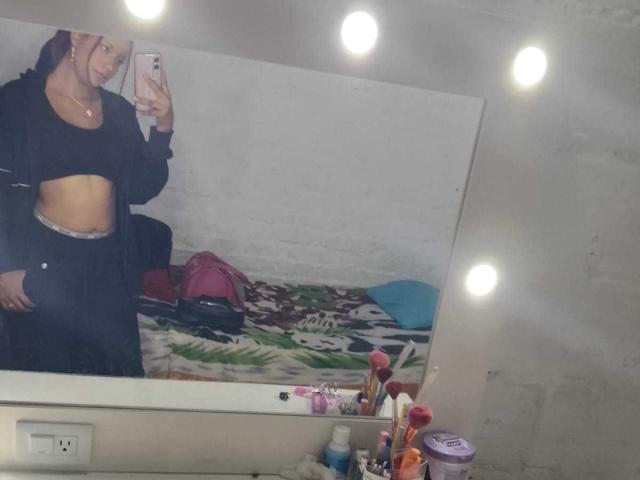 Sofia Hilton mit roten Haaren, schwarzem Crop-Top, schwarzer Jacke und schwarzer Hose, die ihren Bauch in einem Spiegel-Selfie zeigt, Schlafzimmer mit ungemachtem Bett und Toilettenartikeln..
