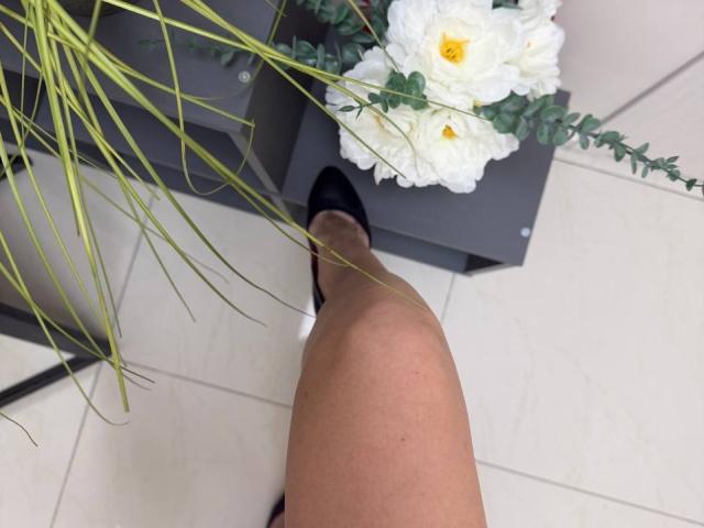 MoonLiza's leg with fair skin, wearing black shoes, next to a gray planter with white roses and green leaves, on a white-tiled floor.