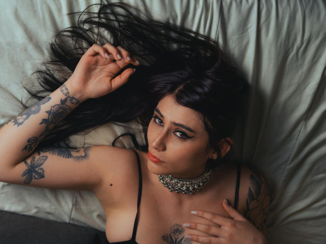 AlynaFerrer with long black hair, tattoos on arms and chest, wearing a black top and jeweled choker, lying on a bed, looking up with a serious expression.