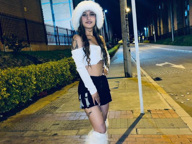 Adissonlee met lang bruin haar, een witte top met open schouder, zwarte shorts, een witte pluizige muts, witte beenwarmers, staand op een stoeprand 's nachts, kijkend naar de camera, lachend..