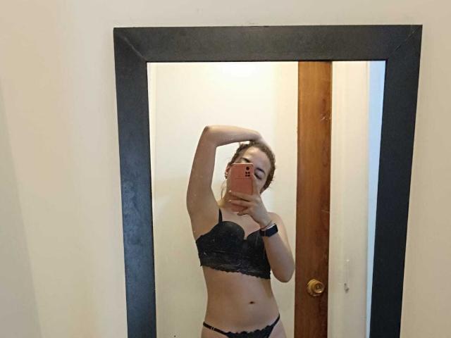 MadisonKoper with her arm up, wearing black lace bra and panties, showing her tits and pussy in a mirror selfie, holding a pink phone, in a room.