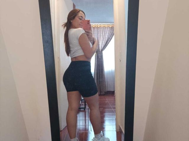 MadisonKoper with a curvy butt in black shorts, white crop top, and white sneakers, taking a mirror selfie in a hallway. Her red hair is down, smiling.