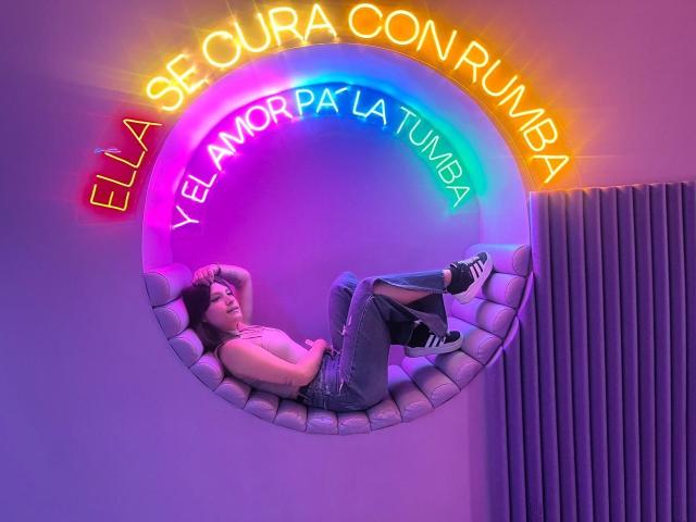 Adissonlee, with bare tits, lies in a circular neon-lit seat, wearing gray jeans and black sneakers, hands behind head, legs up, in a purple and pink neon glow.