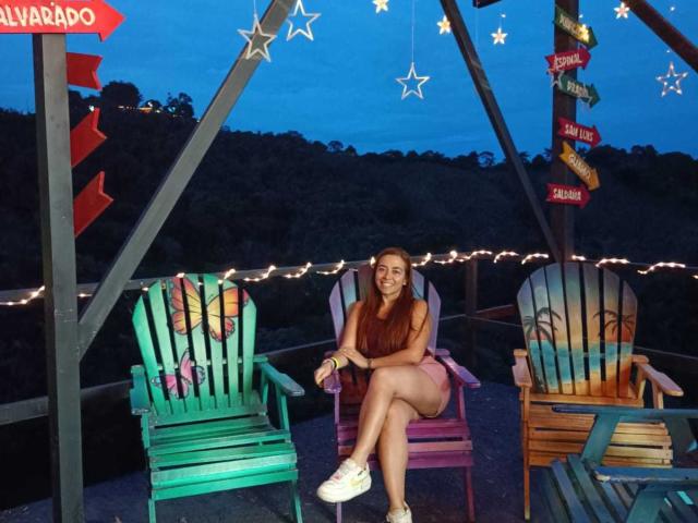 MadisonKoper sitting on a purple wooden chair, legs crossed, smiling, wearing a red dress and white sneakers, with green and blue painted chairs, string lights, and star decorations.