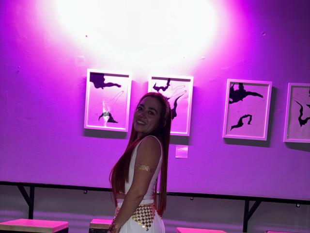 MadisonKoper with long red hair, smiling, wearing a white dress with gold accents, standing in front of four abstract black-and-white framed artworks, illuminated by pink-purple lighting.