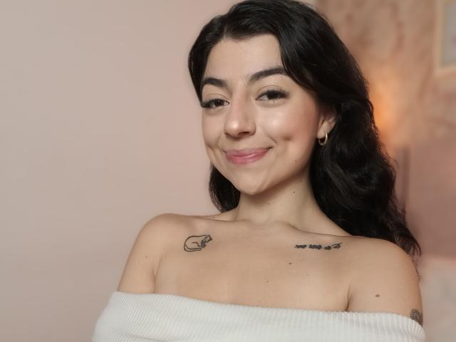 GiuliaAddams, loira, com um sorriso adorável, vestindo uma jaqueta jeans e uma camisa preta, tem uma mão no rosto e está sentada em um café bem iluminado..