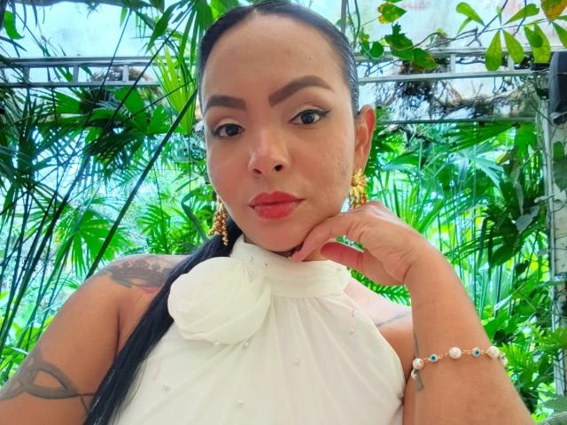 JessicaFiore, com cabelo escuro, blusa de alça branca, brincos de ouro, segurando um copo de bebida de laranja, mostrando seios grandes, tatuagens nos braços e batom vermelho, em pé em um jardim verde exuberante..