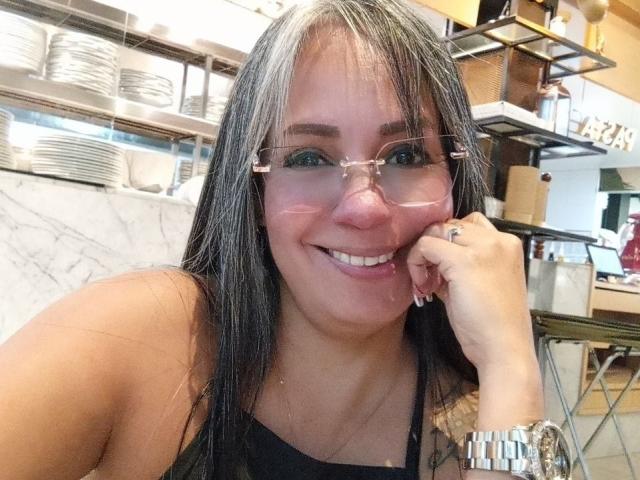 LailaCastillo heeft lang bruin haar, een bril en een roze neus. Ze glimlacht, waarbij haar borsten gedeeltelijk zichtbaar zijn onder een zwarte top, in een restaurant met muren van marmer..