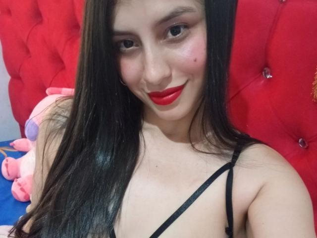 MadissonFontaine with long black hair, red lipstick, and a small mole on her cheek, smiling at the camera, wearing a black spaghetti strap top, with a red tufted background.