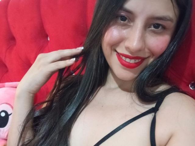 MadissonFontaine with long black hair, red lipstick, and black strap dress, smiling at the camera, showing her small tits, against a red tufted headboard.