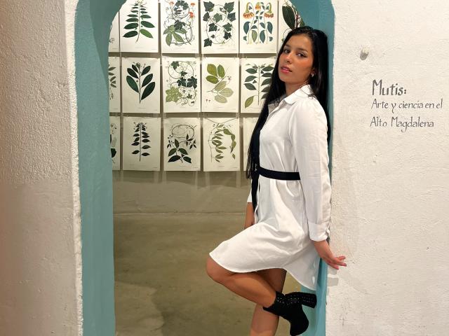MelodyRoss stands in a doorway, wearing a white dress with a black belt, black socks, and black shoes. She has long black hair and a confident look.