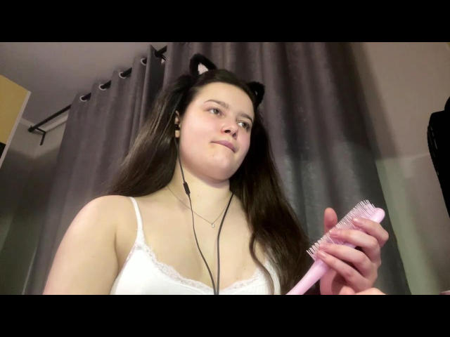 Lashaun Dromgoole com orelhas de gato, segurando uma escova rosa, usando uma camiseta branca, fones de ouvido e com um leve sorriso, cabelo castanho, pele clara, em pé em frente a cortinas cinzas..