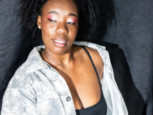 JazaraKoral, com pele escura, sombra rosa, top preto e jaqueta cinza de camuflagem, mostrando seios pequenos e sorrindo levemente, contra um fundo preto..