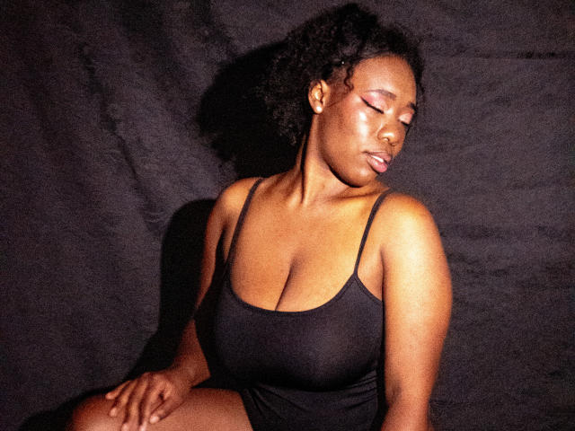JazaraKoral, dark-skinned with curly hair, wearing a black tank top, showing her large boobs, eyes closed, sitting against a black background, looking relaxed and serene.