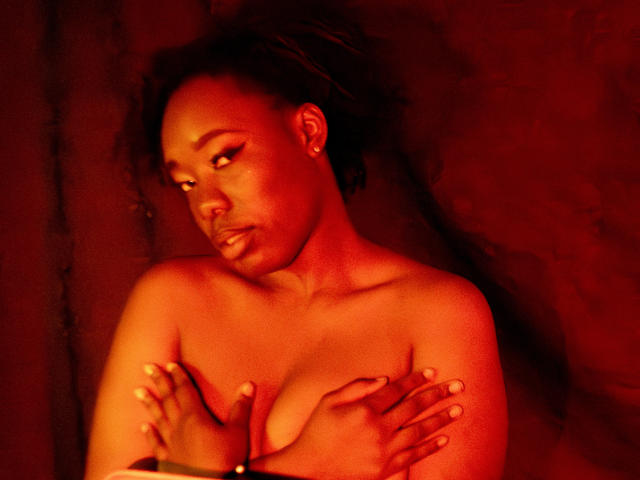 JazaraKoral, a dark-skinned woman with red-lit skin, covering her tits with her hands, has a serious expression, wearing a bracelet, and has her hair in a ponytail.