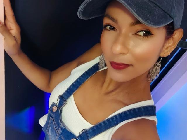 HannaPretty with brown eyes, red lipstick, and a blue cap, wearing denim overalls and a white tank top, showing her tits, smiling, and holding a wall.