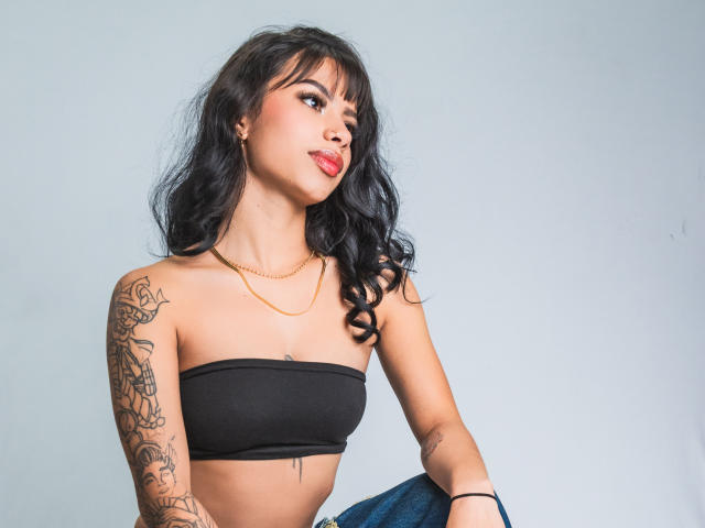 Kazumii, avec ses cheveux noirs ondulés, sa chaîne en or, son haut noir sans bretelles, ses tatouages sur le bras droit, son rouge à lèvres rouge, regardant de côté, tenant un jean bleu. Elle a de petites seins et un visage mignon..
